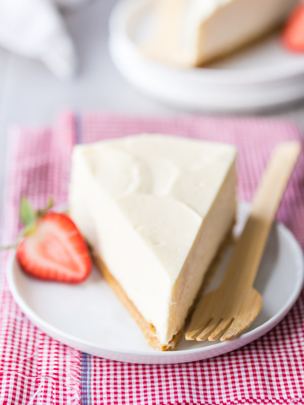 No-Bake Cheesecake slice on a white plate with a fork and half strawberry sitting on a pink/white checked napkin. another slice in view in the background