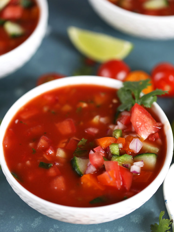 gazpacho in a white bowl garnished with tomato and cilantro. lime wedge in background and peek at two more bowls of gazpacho