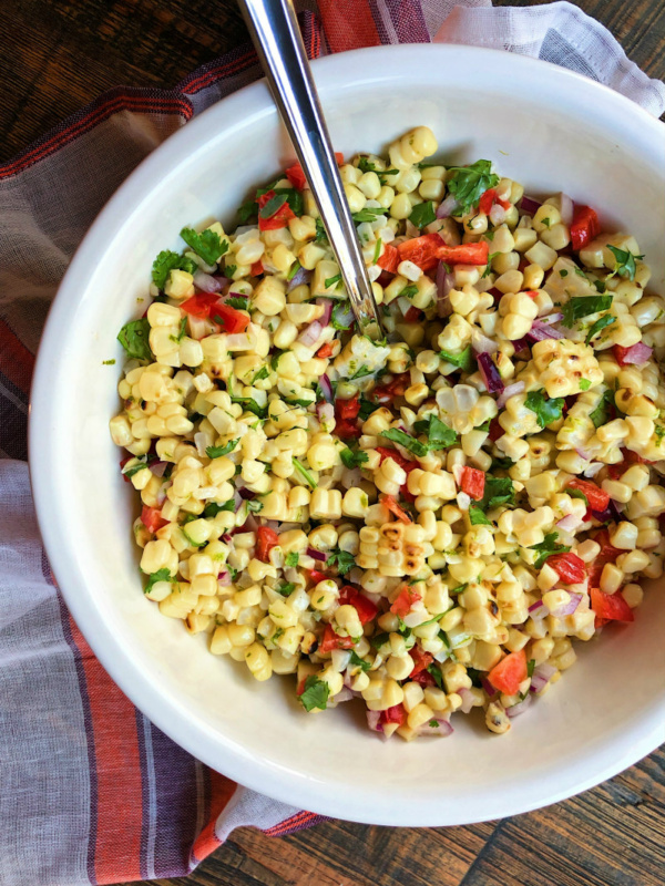 grilled corn salad in a white bowl with a serving spoon on top of an orange, purple and white striped cloth napkin