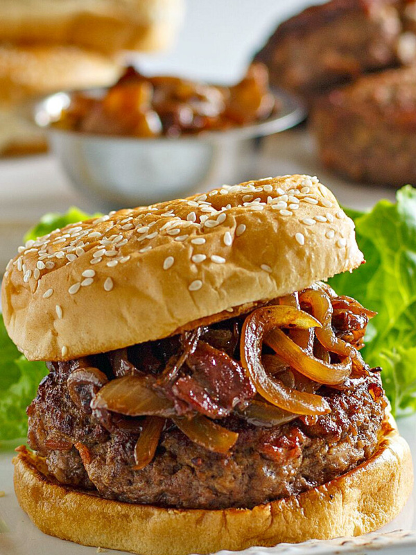 Bacon Burger with Balsamic Caramelized Onions on a white serving platter with lettuce leaf. More buns, burgers and bowl of caramelized onions in the background.