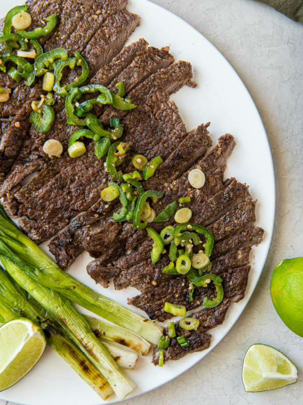 sliced carne asada on a white serving platter topped with jalapeno and with green onions and limes on the side