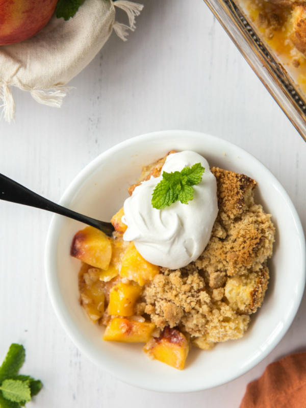 serving of peach cobbler french toast casserole in a white bowl garnished with whipped cream and fresh mint