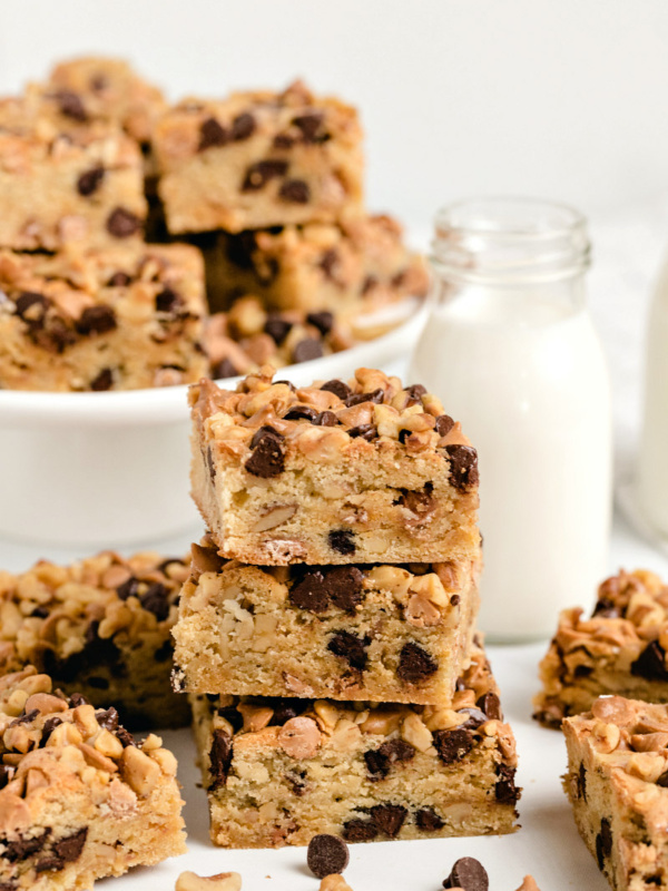 stack of three blondies with more blondies on a white platter in the background and on the sides. Glass of milk too.