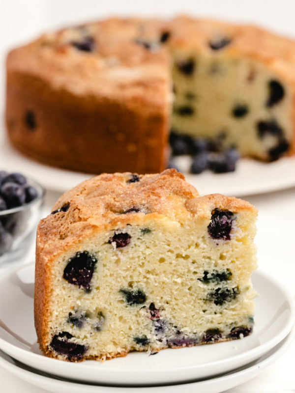 slice of buttermilk blueberry cake on a white plate with the rest of the cake on a white plate in the background