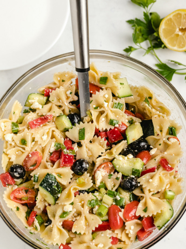 overhead shot of bow tie pasta salad in a glass bowl with a serving spoon and a half lemon and fresh herbs on the side