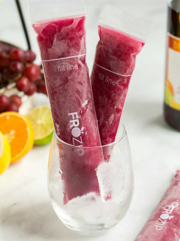 two sangria popsicles standing up in a glass with a bottle of sangria in the background and orange slices, lime slices and grapes in the background too