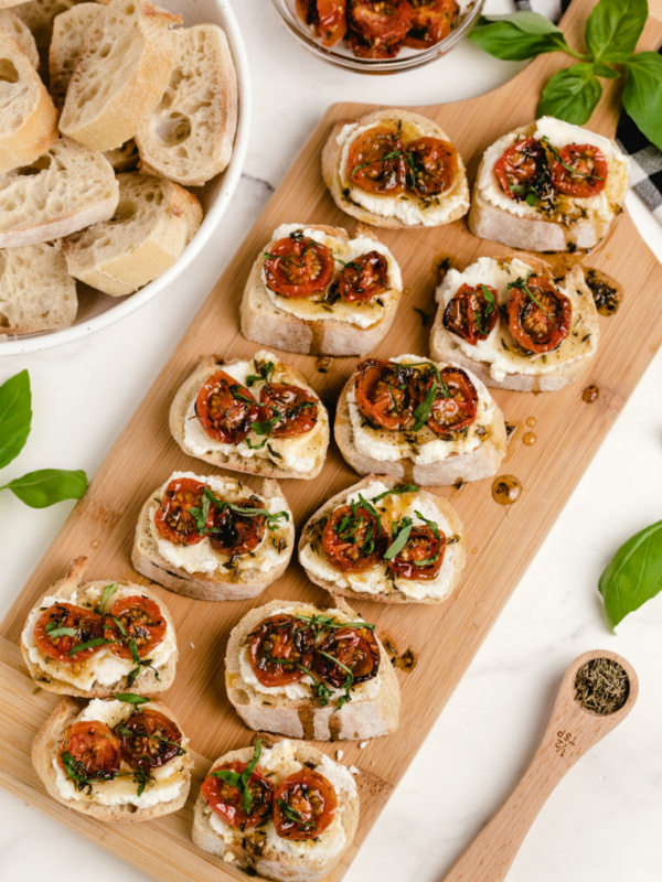 honey tomato bruschetta with ricotta on a wooden board with a bowl of baguette slices and fresh basil on the side with a white and black checked napkin