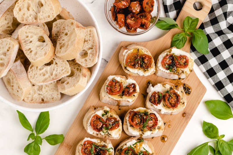 honey tomato bruschetta with ricotta on a wooden board with a bowl of baguette slices and fresh basil on the side with a white and black checked napkin